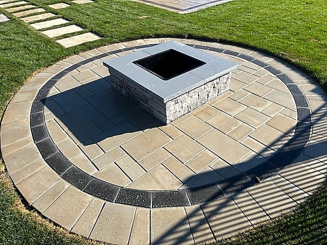 Square stone fire pit at the centre of a circular interlock patio with a charcoal border